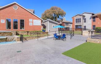 View of an outdoor area featuring a modern swimming pool, lounge chairs, and an umbrella. Surrounding buildings include brick and wood-sided apartments with large windows. Green grass and landscaped areas add to the inviting atmosphere on a sunny day.