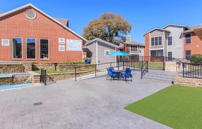 View of an outdoor area featuring a modern swimming pool, lounge chairs, and an umbrella. Surrounding buildings include brick and wood-sided apartments with large windows. Green grass and landscaped areas add to the inviting atmosphere on a sunny day.