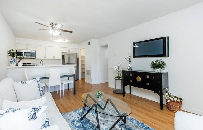A modern living room with a light sofa, a glass coffee table, and a wall-mounted TV. The space features a ceiling fan, wooden flooring, and a decorative cabinet with plants. In the background, thereâs a kitchen area with white cabinetry and a dining table with chairs. Soft blue and white accents enhance the decor.