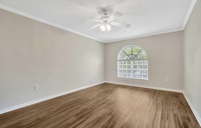 A spacious, empty room featuring a ceiling fan and large arched window allowing natural light. The walls are light-colored, and the floor is made of a warm-toned wood laminate. The room has a serene and clean appearance, ready for personalization or furnishing.