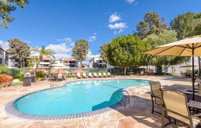 Fusion Warner Center apartments in Woodland Hills pool with tan lounge chairs and patio tables with tan umbrellas on natural stone tile ground.