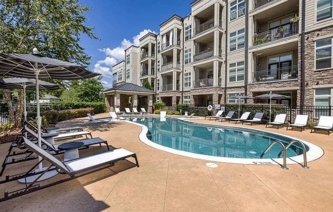 A large swimming pool surrounded by lounge chairs and umbrellas in front of apartment buildings.