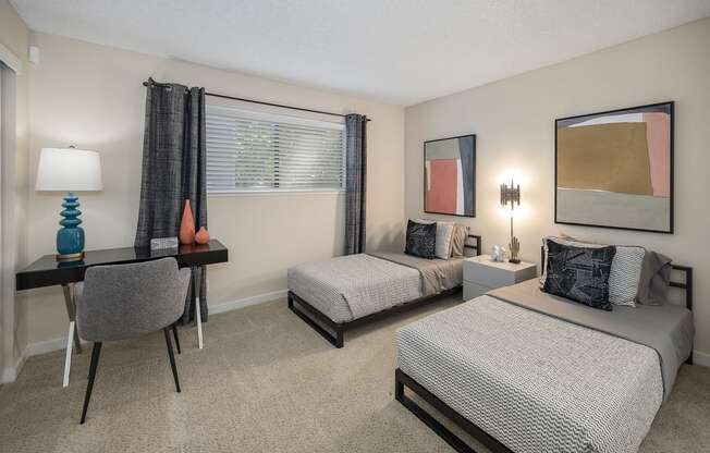 Bedroom with Two Full Beds with headboard up against a wall with decorative wall art above the beds. There is also a Desk with Gray Chair, Carpet through out and Window with Gray Curtains at Silverstone Apartments