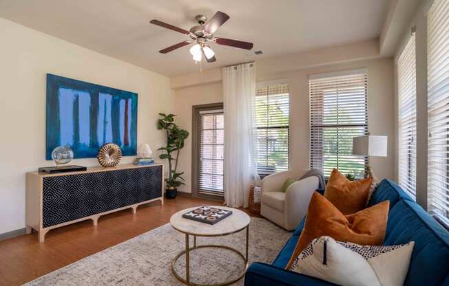 Pet Friendly Apartments in Greenville SC - Velo Verdae - Furnished Living Room with Natural Oak-Style Plank Flooring, Area Rug, Blue Sofa, White Armchair, Marble and Gold Round Coffee Table, Black and Tan TV Monitor, Standing Lamp, Large Windows with Blinds, Ceiling Fan, and House Plant in the Corner