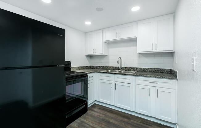 Modern kitchen featuring white cabinetry and granite countertops. Includes a black refrigerator, stove, and sink with a faucet. The space is brightened by recessed lighting and has a neutral color scheme with wood-like flooring.
