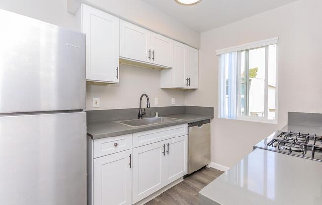 Modern kitchen featuring stainless steel appliances, including a refrigerator, sink, and a gas stove. The cabinets are white with a sleek design, and the countertop is gray. Natural light comes in through a window with vertical blinds. The flooring is a light wood finish, adding to the contemporary feel.
