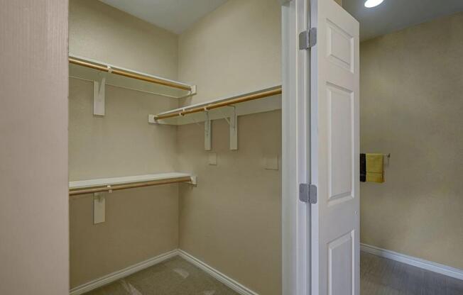 A spacious walk-in closet here at Mission Green featuring neutral-toned walls, multiple built-in hanging rods at varying heights, sturdy white shelving for organized storage, soft carpet flooring, and an open layout that provides convenient access from the adjacent bathroom area.