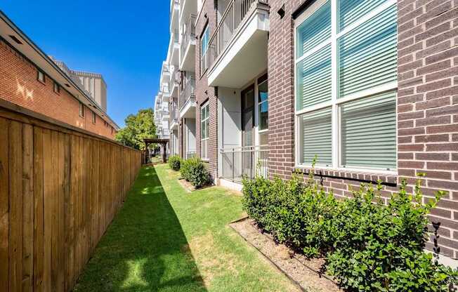 A long, narrow, green lawn separates a brick building from a wooden fence.
