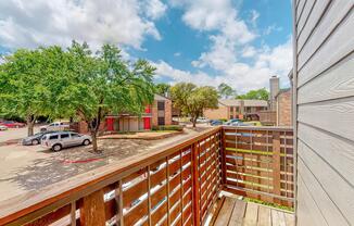 View from a wooden balcony overlooking a parking lot and surrounding trees. The scene features a mix of parked vehicles and several buildings in the background, under a bright sky with scattered clouds, indicating a sunny day.
