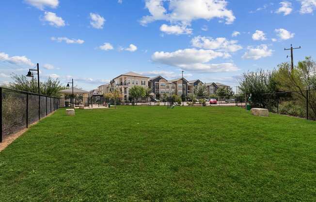 A grassy field with a fence and apartment buildings in the background.