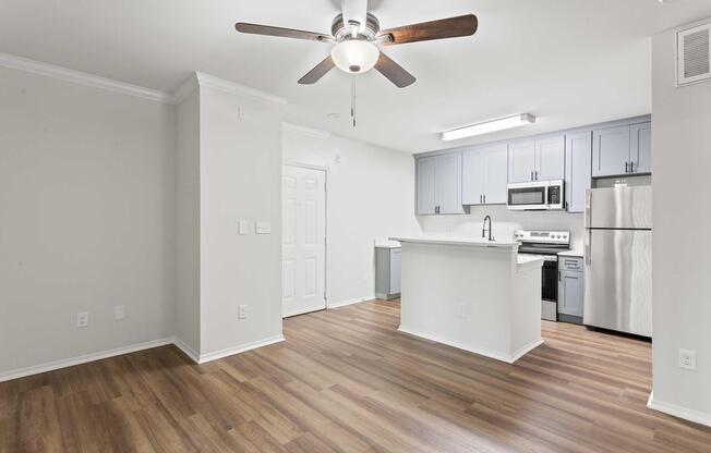 Modern kitchen and living area featuring light gray walls, a ceiling fan, and warm wood-like flooring. The kitchen has blue cabinets, stainless steel appliances, and a large island with seating. Natural light illuminates the space, creating a bright and inviting atmosphere.