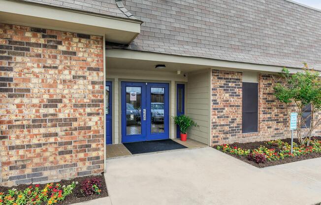Entrance of a building featuring double blue doors, surrounded by a brick facade and decorative flowering plants in front. The path is paved, leading to the entrance, with a planter beside the door.