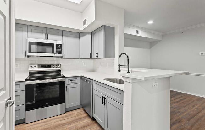 Modern kitchen featuring gray cabinets, stainless steel appliances, a microwave, and a countertop with a sink. The space is well-lit with overhead lighting, and there is an open layout connecting to a living area with hardwood flooring.
