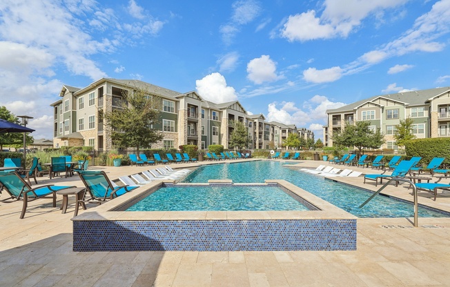 A large swimming pool surrounded by lounge chairs in front of apartment buildings.