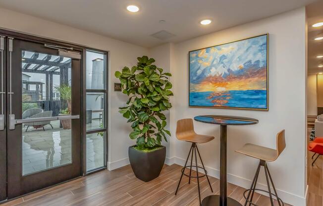 A modern interior corner with a tall potted plant and two stylish bar stools positioned at a small black table. A vibrant painting of a sunset over the ocean adorns the wall, and large glass doors provide a glimpse of an outdoor patio area. The floor features sleek wooden tiles.