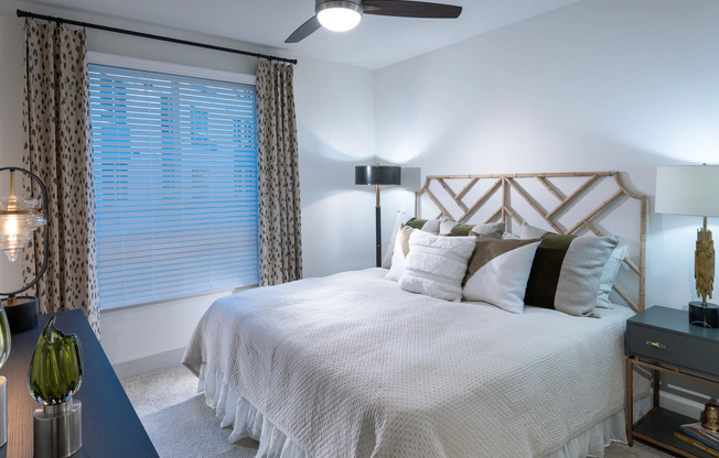 Bedroom with king-sized bed, side table, dresser, large window, ceiling fan, beige carpet, and white walls.