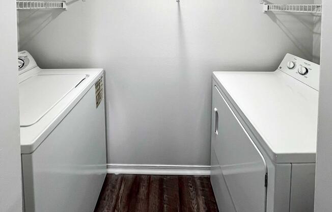 A laundry room featuring a washer on the left and a dryer on the right, both in white. The walls are painted light gray, and there's a shelf above for storage. The floor has dark laminate flooring, creating a contrast with the appliances. The space is well-lit and uncluttered.