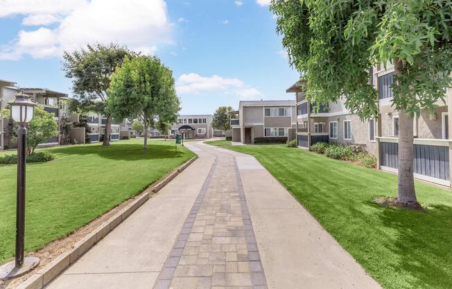 A well-maintained pathway winding through a green landscape, flanked by trees and modern apartment buildings. The sky is bright with fluffy clouds, creating a serene and inviting atmosphere. Well-kept lawns add to the overall appeal of the outdoor space.