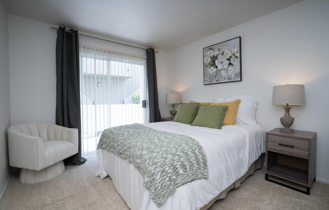 Cozy bedroom featuring a queen-sized bed with white bedding and green accent pillows. A modern armchair is positioned next to the bed. There are two bedside lamps and a floral artwork on the wall. Large sliding glass doors lead to an outdoor area, with sheer curtains allowing natural light.