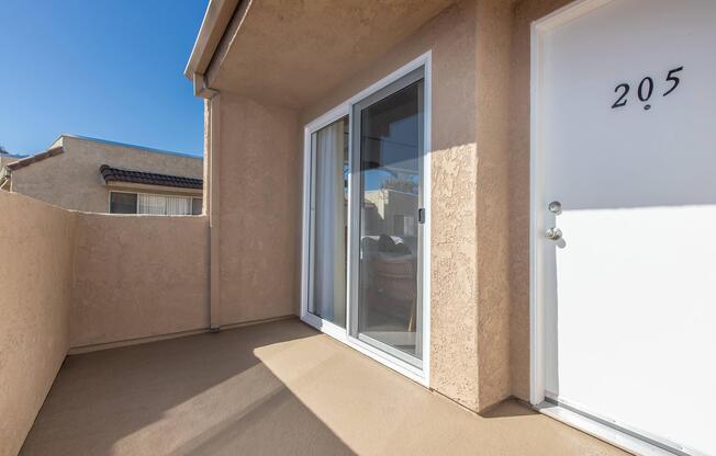 A balcony view from a second-floor unit, featuring a beige exterior wall, sliding glass door, and numbered entrance door labeled "205." The area is well-lit by sunlight, with a clear blue sky in the background.