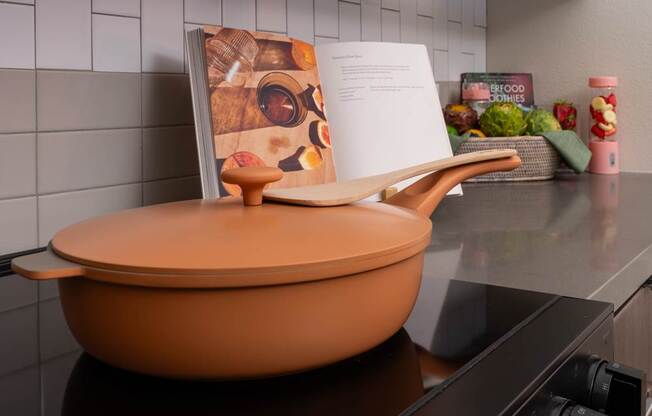 A large orange pot sits on a stove with a recipe book open on the counter.