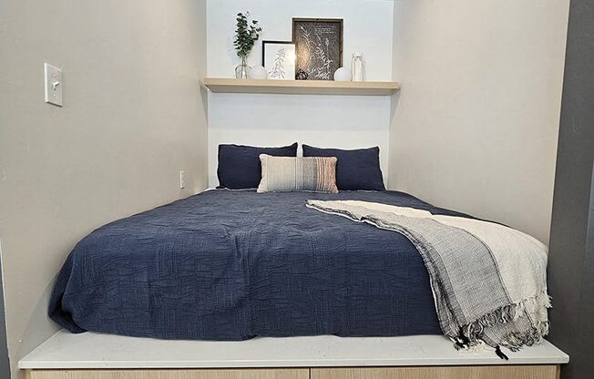 A bed with a blue comforter and pillows is in a room with a shelf above it.
