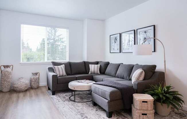 A grey sofa with pillows and a white ottoman in a room with a window.