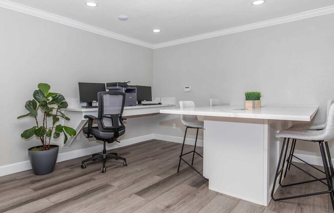 A modern office space featuring a sleek desk with two computer monitors, a comfortable black office chair, and a small plant. A white table with high stools is also present, set against light gray walls and wooden flooring, illuminated by recessed lighting.