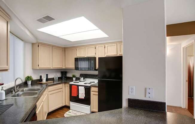 A bright kitchen here at Mirador featuring light wood cabinetry, dark countertops, and a double-basin sink beneath a window. A full appliance package and overhead skylight enhance the space, while ample cabinet storage and a functional layout create a practical setting for everyday cooking and meal preparation.