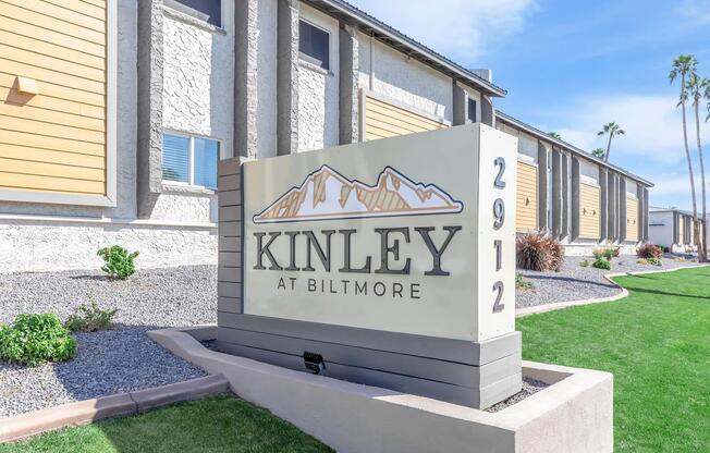 Sign for Kinley at Biltmore, displaying the name "KINLEY" prominently. The sign features mountain imagery and the address "2912." Surrounding landscape includes well-maintained grass and shrubs, with a clear blue sky in the background.
