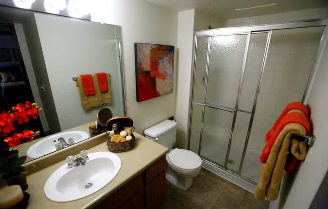 A clean bathroom here at Eagle Ridge featuring a single vanity with integrated sink and mirror, neutral countertop and cabinetry, a toilet positioned beside the vanity, a sliding glass shower enclosure with textured panels, tiled shower walls, soft overhead lighting, and neatly folded towels adding a warm accent.