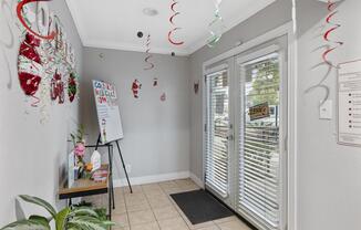 A bright entryway decorated for a festive occasion, featuring a small table with plants and a signboard, light-colored walls, tiled flooring, and double glass doors with vertical blinds. Colorful streamers and ornaments hang from the ceiling, creating a cheerful atmosphere.