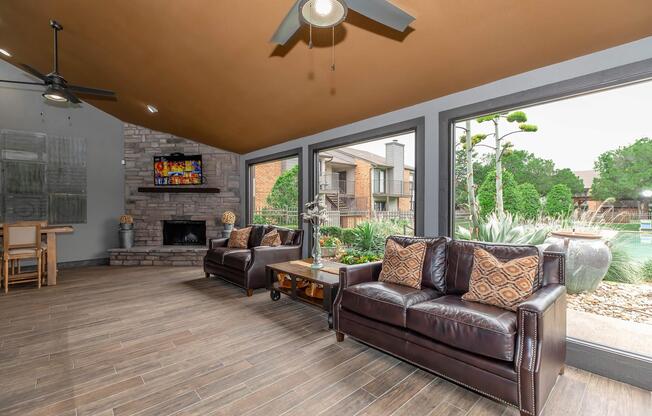 Cozy indoor lounge area featuring two brown leather sofas with decorative pillows, a stone fireplace, and a ceiling fan. Large windows offer a view of an outdoor pool and landscaped garden, creating a bright and inviting atmosphere. A wooden dining table is visible in the background.