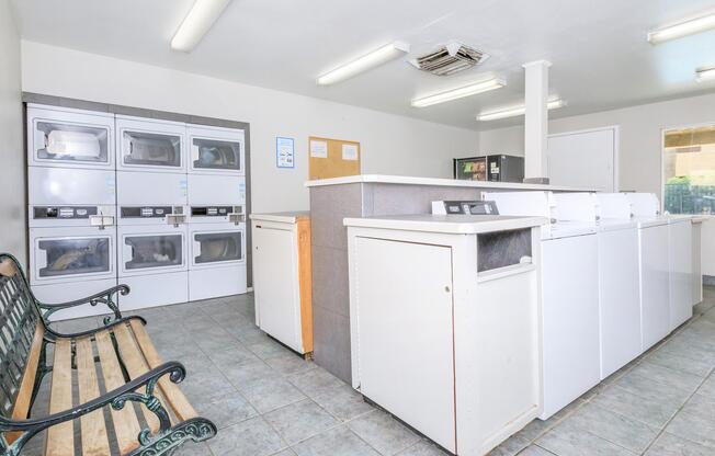 Interior of a laundry room featuring white washing machines and dryers along one wall, a counter with additional machines, and a bench for seating. Bright lighting illuminates the clean, tiled space with a neutral color scheme.