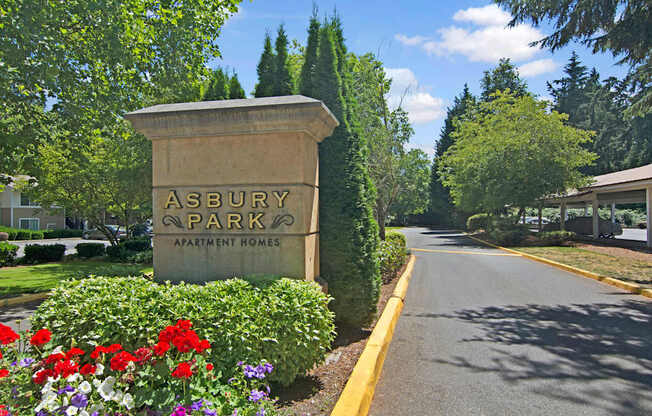 A landscaped community entrance sign here at Asbury Park featuring a stone monument displaying the property name, surrounded by colorful flowers, trimmed hedges, and mature trees, with a paved driveway extending into the community and covered parking visible in the background under a clear blue sky.