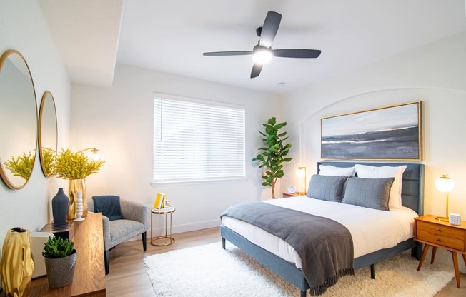 Two-Bedroom Apartments in Broomfield, CO - Terracina - Bedroom with Hardwood-Style Flooring, High Ceiling, Ceiling Fan, Area Rug, Bed, Nightstands with Lamps, Houseplant, Wall Art, Armchair, Dresser, Mirrors, and Large Window with Blinds.