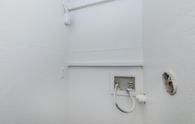 A corner of a room featuring a white wall with a small panel that has two connected pipes. There is an electrical outlet nearby and a small hole in the wall. The overall atmosphere is minimalist and somewhat unfinished, indicating possible plumbing or installation work.