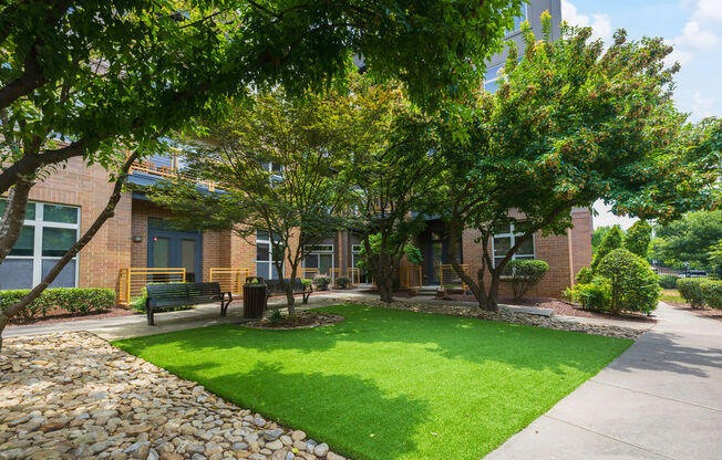 a green lawn in front of a brick building with trees and a bench