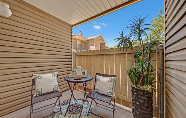 A cozy private patio here at Northridge Court featuring two woven outdoor chairs with patterned accent pillows, a small round bistro table with decorative accents, and a patterned outdoor rug, enclosed by neutral siding and a wooden privacy fence with a tall potted plant adding greenery beneath a bright blue sky.