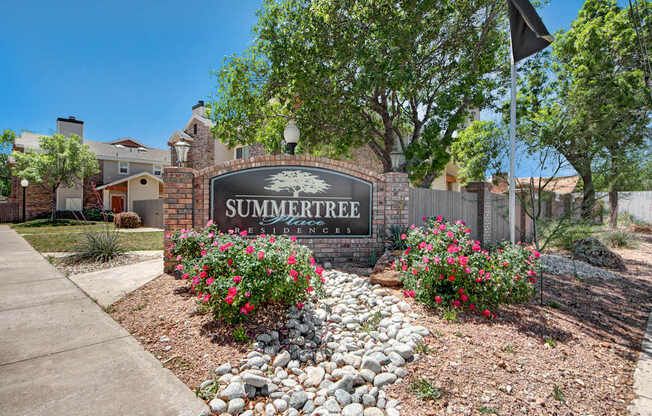 A welcoming monument sign here at Summertree Place showcasing a brick-framed community sign surrounded by blooming pink flowers, decorative stone landscaping, mature trees, and nearby apartment buildings, creating a polished and inviting entrance along a sunny residential sidewalk.
