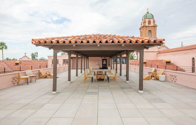A patio with a roof and chairs is in front of a building with a tower.