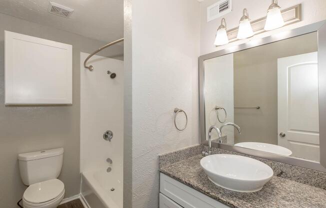 A modern bathroom featuring a white toilet, a shower with a glass door, and a granite countertop with a stylish sink. The walls are painted in a light gray color, and there is a large mirror above the sink with three light fixtures. The flooring appears to be a wood-like laminate.