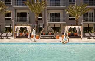 A woman is sitting on the edge of a pool in front of a building with palm trees.