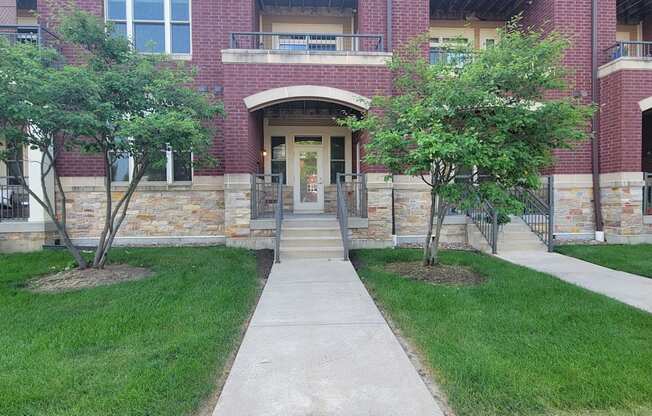 A welcoming building entrance here at Van Buren East with red brick exterior, green trees, and a clean concrete walkway. Charming landscaping and a covered entry create a warm and inviting residential approach.