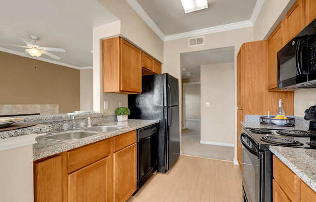 A warm and functional kitchen here at Mission Green featuring wood-style cabinetry, speckled countertops, a double-basin sink, and full-size appliances arranged along a galley layout, with durable flooring, overhead lighting, and an open view toward the living area that supports easy cooking and everyday use.