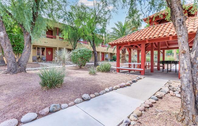 A scenic view of a landscaped courtyard featuring a stone pathway leading to a red-roofed gazebo. Surrounding the gazebo are shrubs, plants, and palm trees. In the background, two-story buildings are visible, creating a serene, inviting outdoor space.