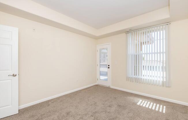 A spacious, empty room featuring light beige walls, plush carpet, and a door leading outside. It has vertical blinds on a large window, allowing natural light to enter. The ceiling is plain, and one wall is unobstructed, creating a bright, airy atmosphere.