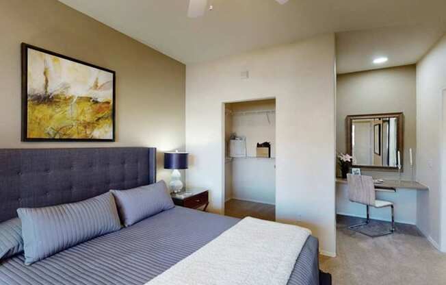 A comfortable bedroom here at Dobson Towne Center featuring plush carpet, neutral walls, and a ceiling fan with light. A spacious bed with an upholstered headboard anchors the room, while an open closet and built-in vanity area add functionality. An adjoining bathroom enhances privacy and everyday convenience.