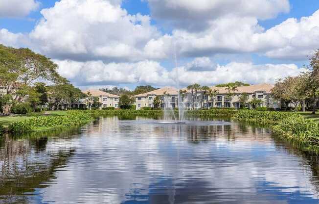 A serene lake with a fountain in the middle surrounded by greenery and houses in the background.
