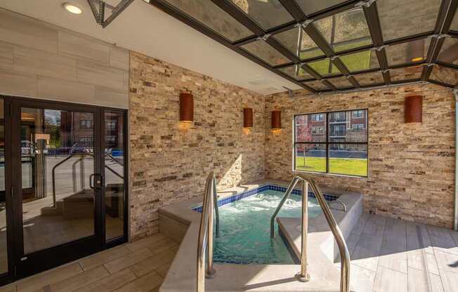 A relaxing spa room here at The Park featuring a bubbling hot tub, stone accent walls, large windows, glass garage-style door, and warm lighting, creating a luxurious and tranquil retreat space.